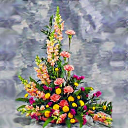 Tall triangular arrangement with pink carnations, snapdragons, and yellow and purple flowers on a gray background