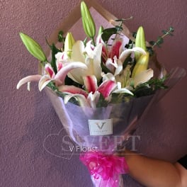 Hand-tied bouquet of pink and white lilies with buds, wrapped in clear paper with a bright pink ribbon bow