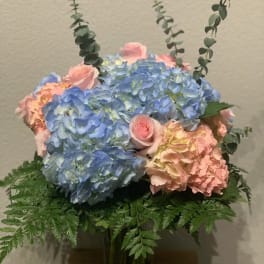 Pastel blue and pink hydrangeas with pink roses and eucalyptus in a clear glass vase with a fern collar