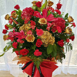 Tall standing arrangement of red roses, lilies, carnations and gold-toned blooms wrapped in red and gold paper.