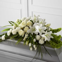 White roses, lilies, and tulips arranged in a long spray on top of a gray casket