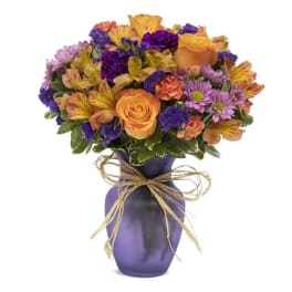 Mixed bouquet of orange roses, purple daisies, and alstroemeria in a purple vase