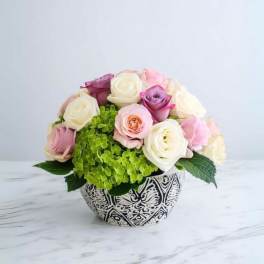 Low arrangement of white, pink, and lavender roses with green hydrangea in a patterned bowl