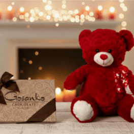Red teddy bear and decorative chocolate box on a wooden surface with soft candlelight in the background.