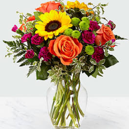 Bouquet of sunflowers, orange roses, and magenta flowers in a clear glass vase