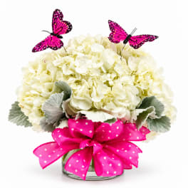 White hydrangea arrangement in a glass vase with pink ribbon and butterfly picks