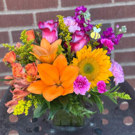 Colorful bouquet with orange lilies, yellow sunflower, and pink roses in a square vase