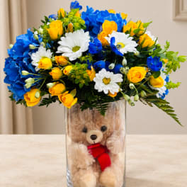 Bouquet of yellow roses, blue carnations, and white daisies in a glass vase with a teddy bear