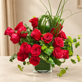 Red roses arranged in a clear square vase with tall grass accents