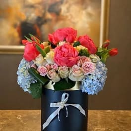 Pink and blue floral arrangement in a dark round box with a ribbon
