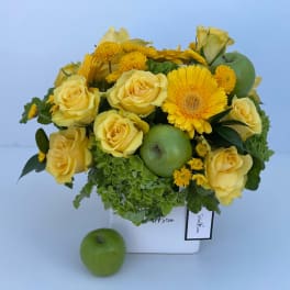 Yellow roses and gerbera daisies arranged with green apples in a white vase.