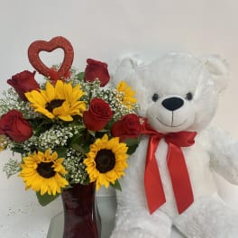 Red roses and sunflowers in a vase beside a white teddy bear with a red bow