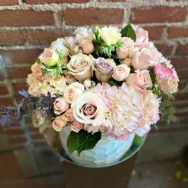 Round arrangement of blush roses and hydrangeas in a white vase against a brick wall.