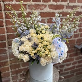 Tall hatbox arrangement of ivory roses and blue flowers with baby boy ribbon accents