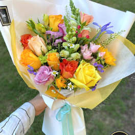 Hand-tied bouquet of yellow, orange, and pink roses with mixed blooms in white wrap.