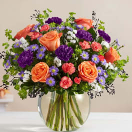 Mixed bouquet of orange roses, purple carnations, and small lavender daisies in a glass vase