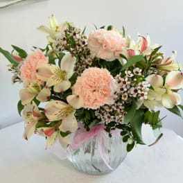 Peach carnations and pale lilies in a clear glass vase with a pink ribbon