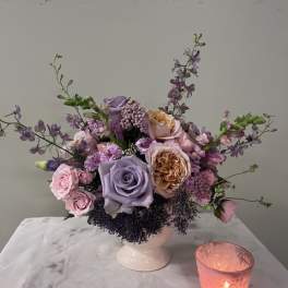 Lavender and pink rose arrangement in a white vase with a lit candle beside it.