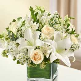 White lilies, cream roses, and small white flowers arranged in a square glass vase