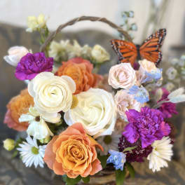 Basket arrangement of roses, daisies, and purple blooms with a butterfly accent