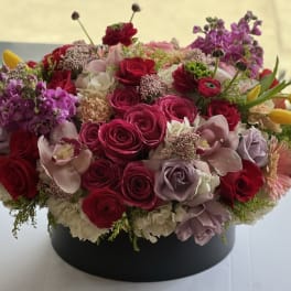 Round arrangement of pink and red roses with purple blooms in a black box