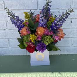 Colorful bouquet of purple, peach, and pink flowers in a white box