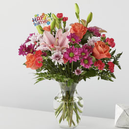 Mixed bouquet of pink lilies, roses, and daisies in a glass vase with a birthday pick