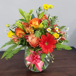 Bright mixed bouquet of orange roses, red gerbera daisy and yellow blooms in a round glass vase with pink bow