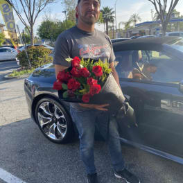 Man holding a wrapped bouquet of red roses beside a dark car in a parking lot