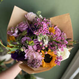 Handheld bouquet of pink, purple, white, and yellow flowers wrapped in brown paper