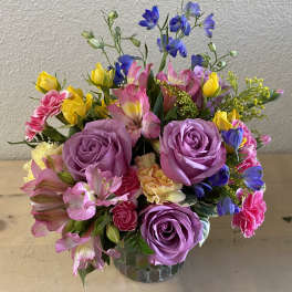 Mixed bouquet of purple roses, pink alstroemeria, and yellow blooms in a glass vase