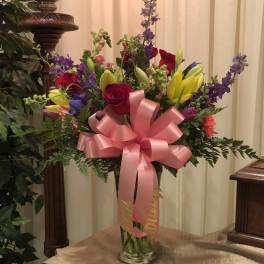 Colorful bouquet of roses, lilies, and purple flowers in a glass vase with a pink bow