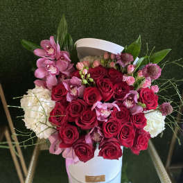 Round white hat box filled with red roses, pink orchids, and white blooms on a gold stand