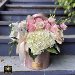 Round hatbox arrangement of pink and cream roses with white hydrangeas and a satin bow