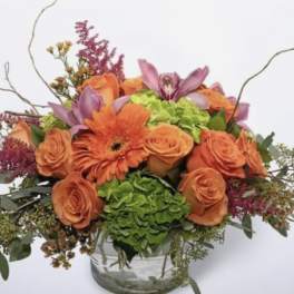 Orange roses and gerbera daisies in a glass vase with purple accents