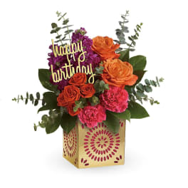 Bright orange roses, pink carnations, and purple blooms in a gold cube vase with a happy birthday pick