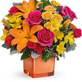 Bright arrangement of orange lilies, pink roses, and yellow blooms in an orange cube vase