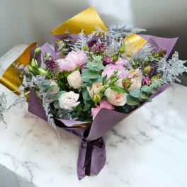 Hand-tied bouquet of pink and cream flowers with purple accents wrapped in purple and gold paper