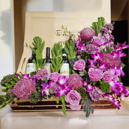 Pink and purple floral arrangement with wine bottles in a wooden box