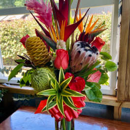 Tall tropical arrangement with red and pink flowers and protea in a clear glass vase with a red bow.