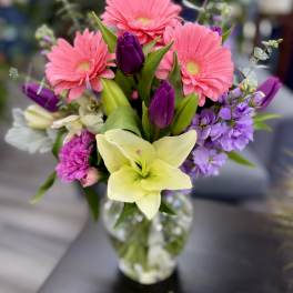 Mixed bouquet of pink gerbera daisies, purple tulips, and a pale lily in a glass vase