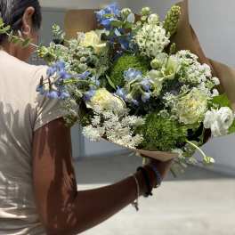 Handheld bouquet of blue, white, and pale yellow flowers wrapped in brown paper