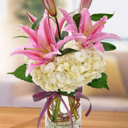 Pink lilies and white hydrangeas in a clear glass vase with a purple ribbon