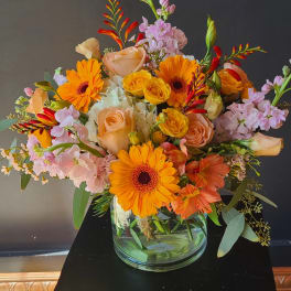 Mixed bouquet of orange gerbera daisies, peach roses, and pink blooms in a glass vase