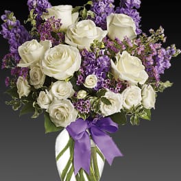 White roses and purple flowers arranged in a glass vase with a purple ribbon