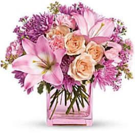 Pink and peach lilies, roses, and mums arranged in a pink glass cube vase