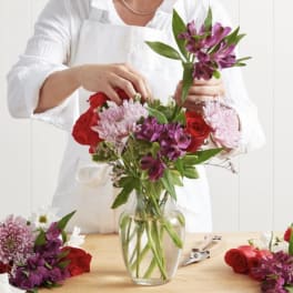 Person arranging a bouquet of red, pink, and purple flowers in a glass vase