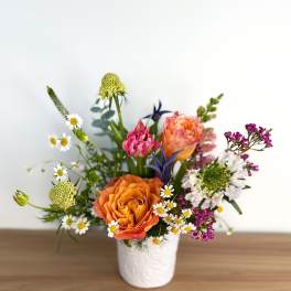 Colorful arrangement of orange roses, pink blooms, and white daisies in a white ceramic vase