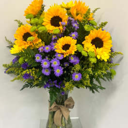 Tall arrangement of yellow sunflowers and purple asters in a clear vase with a burlap bow