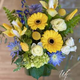 Bright mixed arrangement with yellow gerbera daisies, white blooms, and blue flowers in a green glass vase